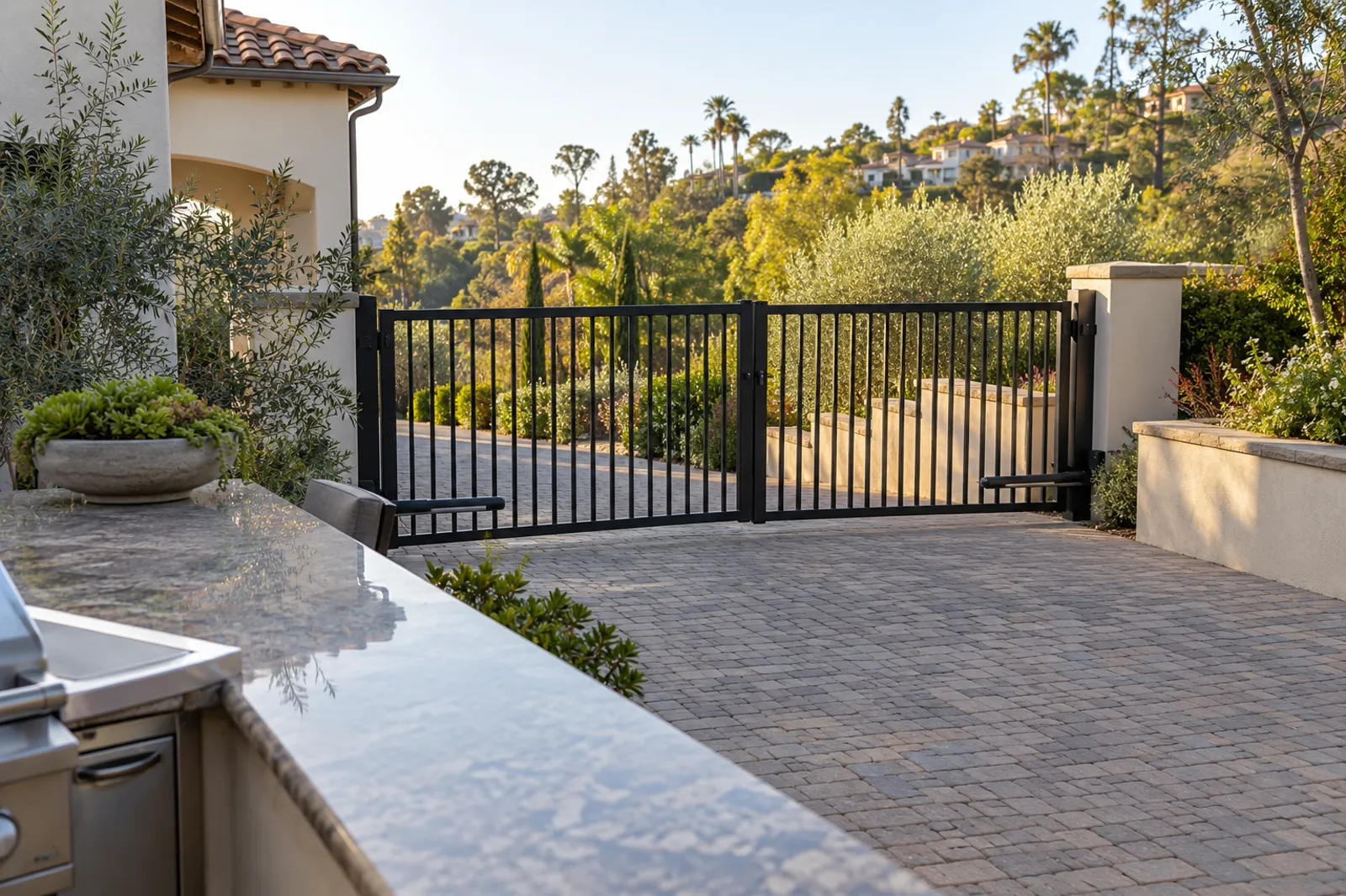 Black steel dual swing driveway gate with vertical pickets across a paver drive on a hillside home, outdoor kitchen counter in the foreground