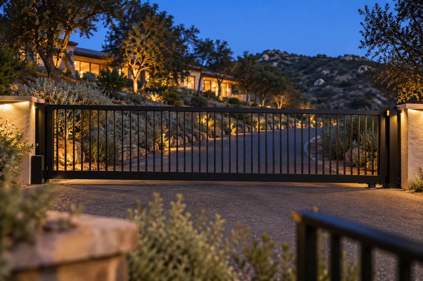 Black steel cantilever slide gate across a sloped drive below a hillside home, landscape lights at dusk